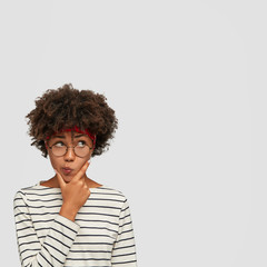Vertical shot of confused hesitant woman holds chin and purses lips, looks in disbelief upwards,...