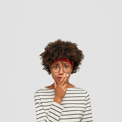 Indoor shot of worried black confused woman keeps hand on chin, looks with puzzled expression at camera, wears round spectacles, stands over white wall, blank copy space above. Nervous clueless girl