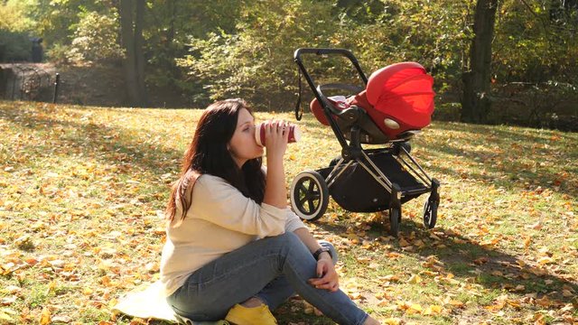 Mother sit on yellow lawn in autumn park drinking coffee while baby sleep in stroller