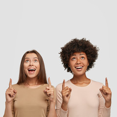 Photo of satisfied mixed race women with joyful expression indicates upwards with both index...