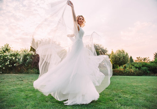happy young blonde girl in an elegant amazing long white wedding light and flying dress whirls and smiles to the camera, raising up a part of the dress. A bride in a green garden. art photo
