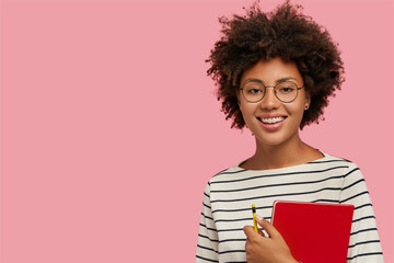 Studio shot of pretty dark skinned girl with gentle smile, prepares for classes, carries red notepad and pencil, wears round glasses, isolated over pink background, free space for your advertisement