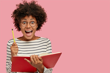 Overjoyed black woman holds textbook and pencil, exclaims with amazement, creats bestseller, wears round glasses, poses against pink wall with copy space on right side. Well, I get good idea for poem - obrazy, fototapety, plakaty