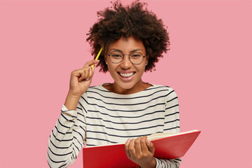 Image of beautiful cheerful girl with toothy smile writes notes in notebook with pencil, dressed in...