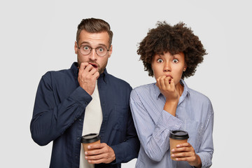 Two worried mixed race students bite finger nails, wait for results, hold disposable cup with...