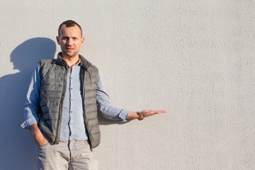 handsome young guy in gray jeans, blue denim shirt and waistcoat stands near the bright wall, copy...