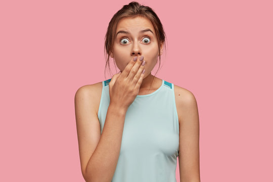 Shocked green eyed beautiful young woman with amazed look, covers mouth with hand, being speechless, dressed in casual clothes, isolated over pink studio background. People and reaction concept.