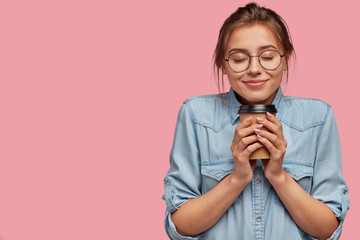 Pleased lovely Caucasian woman holds aromatic beverage, drinks cappuccino or coffee, feels warm, closes eyes from pleasure, has charming smile, wears denim shirt, isolated over pink background