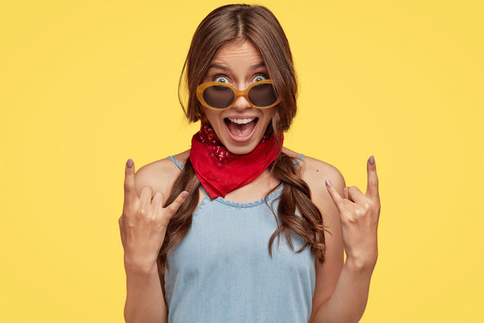 Headshot of provocative cool female hipster shows rock n roll hand gesture, exclaims something loudly, wears fashionable bandana, trendy sunglasses, poses against yellow background. Its so nice!