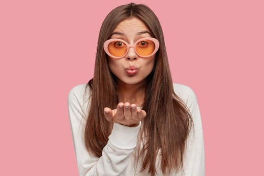 Indoor shot of pleasant looking young brunette Caucasian woman in trendy sunglasses, makes air kiss on distance, says goodbye, expresses love, poses against pink background. Body language concept