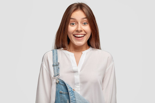 Joyful teenage girl has toothy smile, shows white teeth, happy to spend free time with friends, wears elegant shirt and denim overalls, stands indoor, being very glad. Smiling brunette European woman - Powered by Adobe
