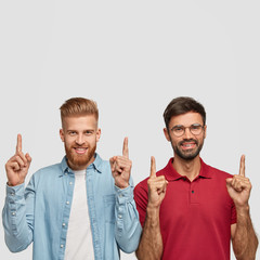 Positive smiling men with thick beards, trendy haircuts, show direction while point upwards, model...