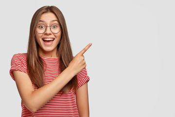 Horizontal shot of cheerful dark haired woman feels overjoyed, dressed casually, demonstrates copy...