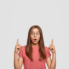 Photo of astonished brunette young woman with surprised expression, points with both index fingers upwards, hears something terrible upstairs, reacts on sudden news, involved in advertisement
