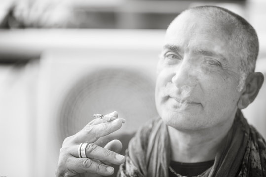 A 50y man is smoking a cigarette. Black and white. The fan coil of an air-conditioner is on the background.