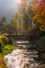 Fototapeta na wymiar Picturesque view of a wooden bridge over a river or a creek with waterfalls in autumn with natural lens flare. Autumn river landscape, Samobor near Zagreb, Croatia