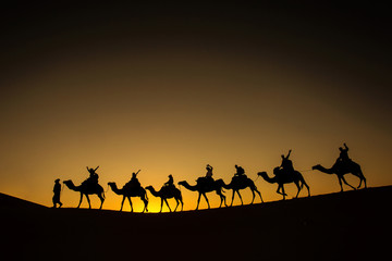 Sillhouette of camel caravan with happy peopple going through the desert at sunset with blue and red sky