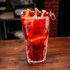 a glass of beautiful bright cocktail on a wooden surface decorated with fruits, berries, leaf of mint and basil at bar counter background