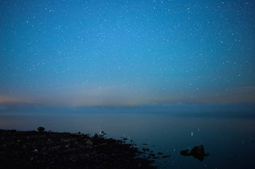 Starry night at rocky lake shore. Fog over water in horizon.