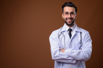 Young handsome bearded Iranian man doctor against brown backgrou