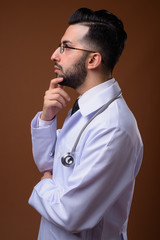 Young handsome bearded Iranian man doctor against brown backgrou