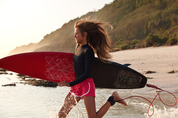 View of active sporty woman runs on water in warm ocean, during windy weather, carries surfboard...