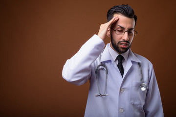 Young handsome bearded Iranian man doctor against brown backgrou