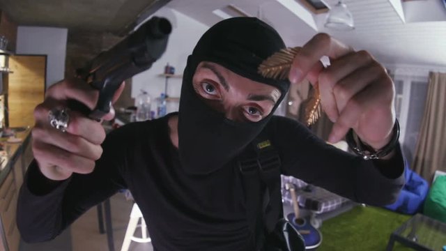 Caucasian bandit in black balaclava showing gold earrings into hidden camera. Happy robber with gun dancing, showing obscene gesture. Indoors.