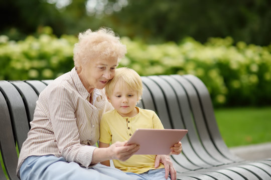 Beautiful granny and her little grandchild together in park. Grandma and grandson seating on the bench and using tablet