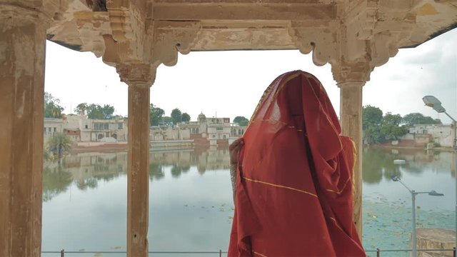 A movement shot of an attractive traditional woman or lady in a colorful sari and jewelry walk out of the house or palace to see the view of a beautiful lake and an old and cultural Indian village. 