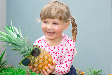 The little beautiful girl cheerfully smiles, laughs and holds in hands pineapple in glasses. In the garden outdoors.
