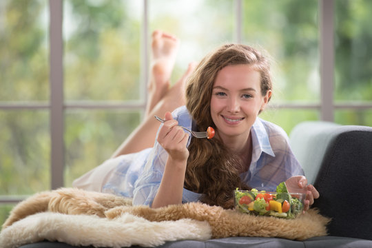 Healthy young woman lying on a couch holding a salad bowl looking relaxed and comfortable