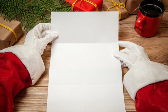 Santa Claus holding letter on wooden table with gift boxes and Christmas tree and cup of hot coffee or tea.  mockup blank