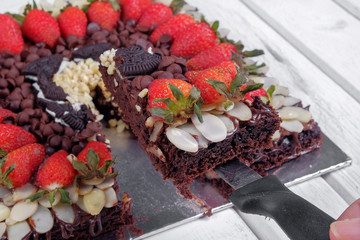 A man's hand with a kitchen knife above a brownie cake with strawberries on a wooden table