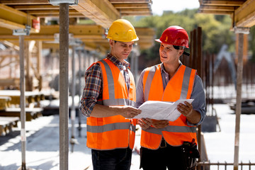 Two civil engineers dressed in orange work vests and helmets explore construction documentation on...
