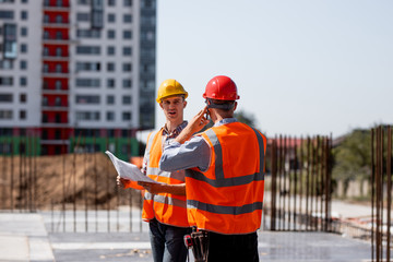 Two structural engineers dressed in shirts, orange work vests and helmets explore construction...