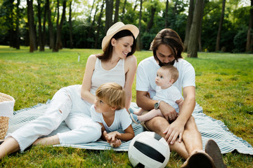 Cheerful parents with two kids dressed in white clothes is sitting on a striped blanket on the lawn on a warm sunny day in the park.