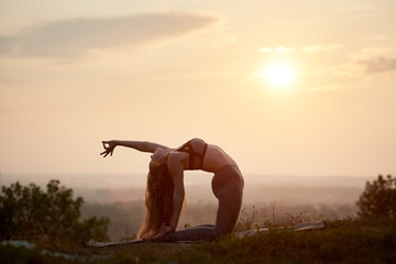 Silhouette of attractive slender fit flexible long-haired young woman doing complicated gymnastic yoga exercises outdoors on grassy hill on background of bright pink misty sky at sunset.
