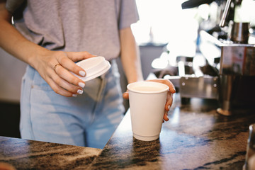 A slim young beautiful girl,dressed in casual outfit, gently puts the lid on the coffeein a cozy coffee shop.