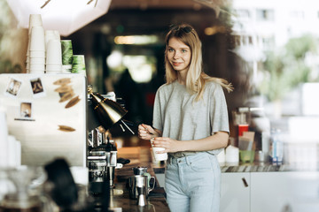 A young cute thin blonde with long hair,dressed in casual outfit,is cooking coffee in a modern coffee shop. Process of making coffee is shown.