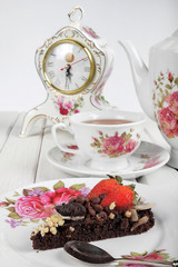 Piece of delicious homemade brownie cake with strawberries , vintage table clock and tea set on white table. Selective focus