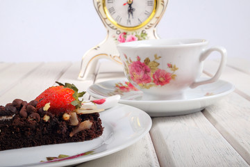 Piece of delicious homemade brownie cake with strawberries , vintage table clock and tea set on white table. Selective focus