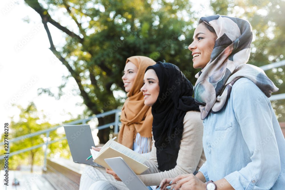 Wall mural photo of three islamic women wearing headscarfs, resting in green park and using laptop