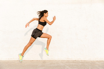 Full length image of athletic woman 20s in sportswear working out and running, along wall