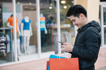 Asian man smiling , using cellphone and holding shopping bags  at the shopping mall.