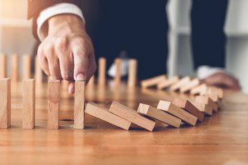 Close up finger businessman stopping wooden block from falling in the line of domino with risk...