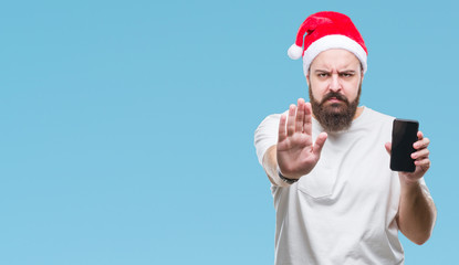 Young caucasian man wearing christmas hat showing smartphone screen over isolated background with open hand doing stop sign with serious and confident expression, defense gesture