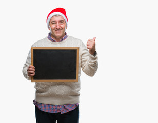 Handsome senior man wearing christmas hat and holding blackboard over isolated background pointing and showing with thumb up to the side with happy face smiling