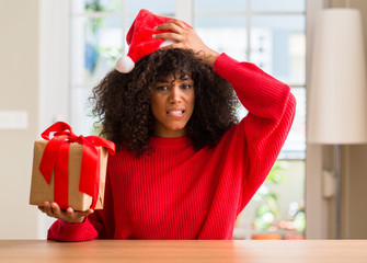 African american woman holding present wearing christmas red hat stressed with hand on head, shocked with shame and surprise face, angry and frustrated. Fear and upset for mistake.