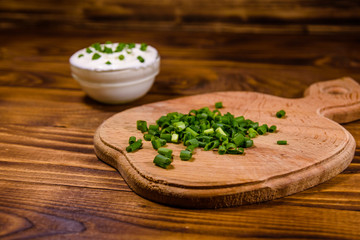Glass bowl with sour cream and cutting board with chopped green onion on wooden table
